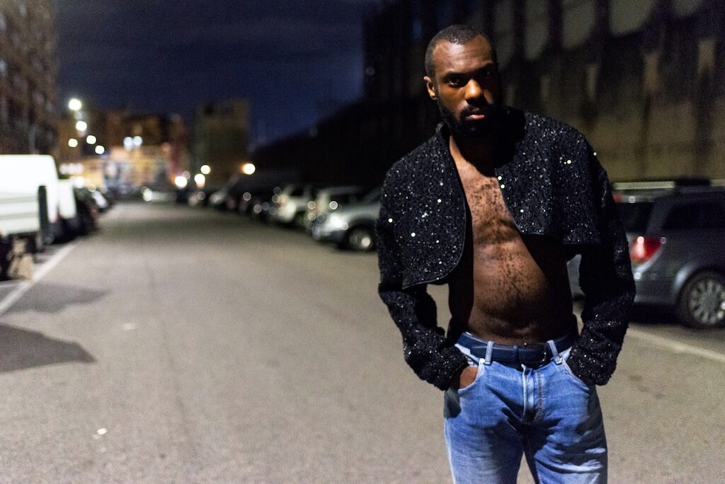 Matteo B in blue jeans and a black, short, sparkly jacket standing in the dark in the street