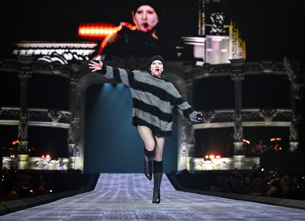 Lady Gaga in striped black and grey jumper and beanie, singing)