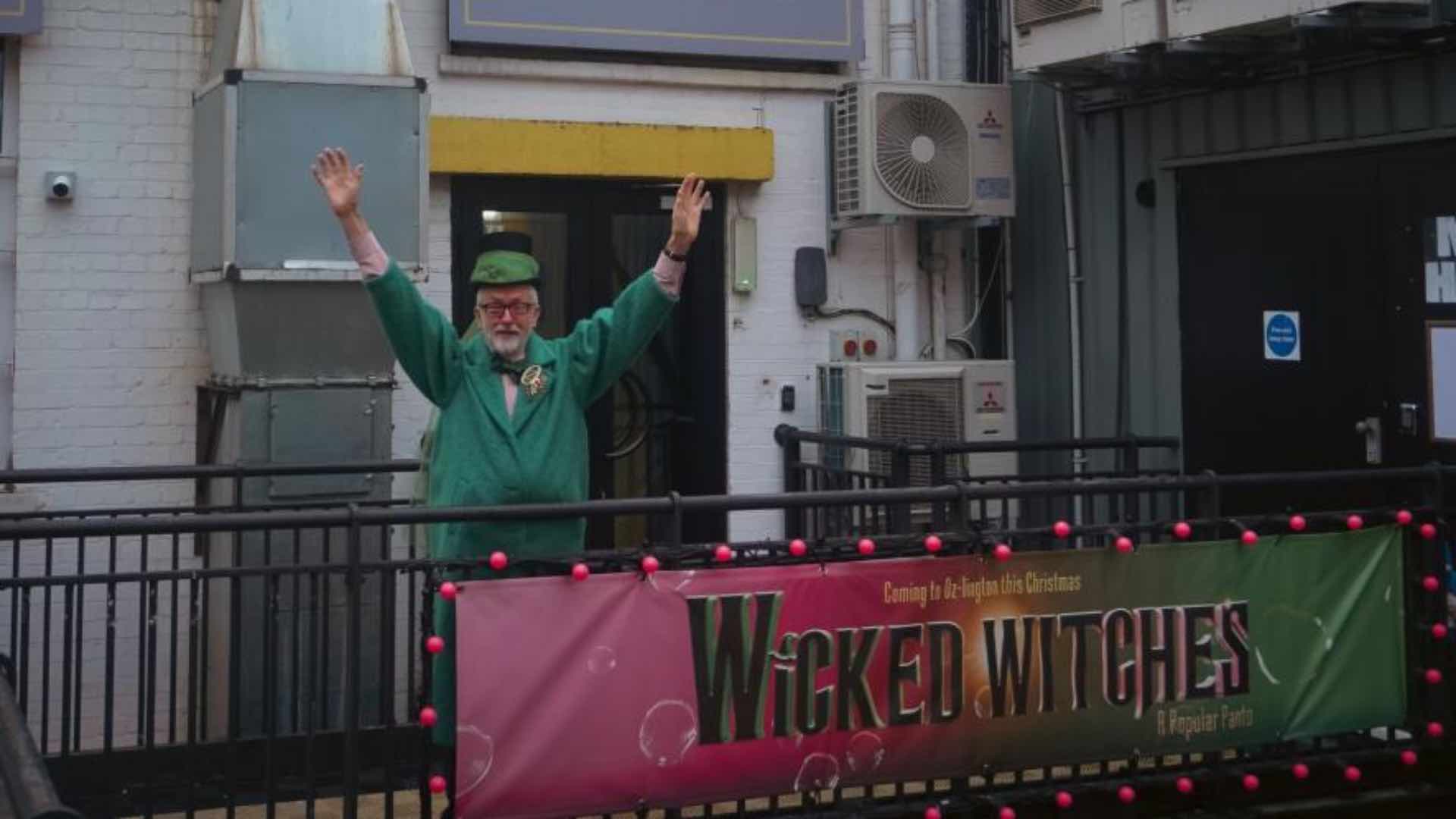 Jeremy Corbyn dressed in green behind the Wicked Witches: A Popular Panto banner