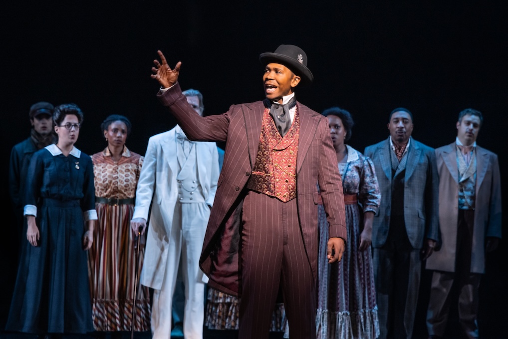 Man with his hand raised whilst talking passionatley with the cast of Ragtime behind him