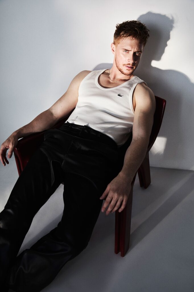Max Parker in a white vest and black trousers sitting on a chair against a white background