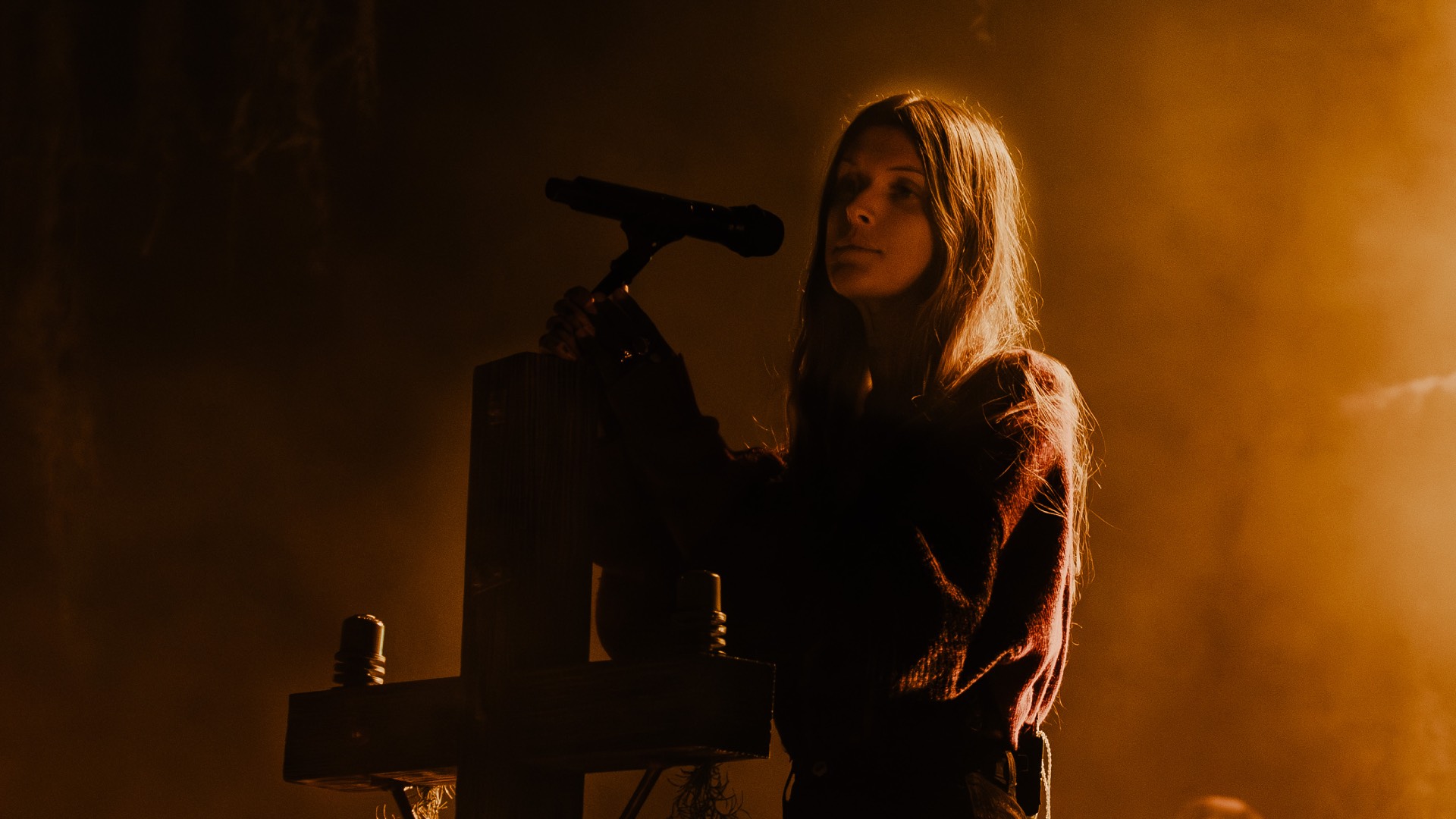 Ethel Cain in black standing on stage, very dark, crucifix mic stand