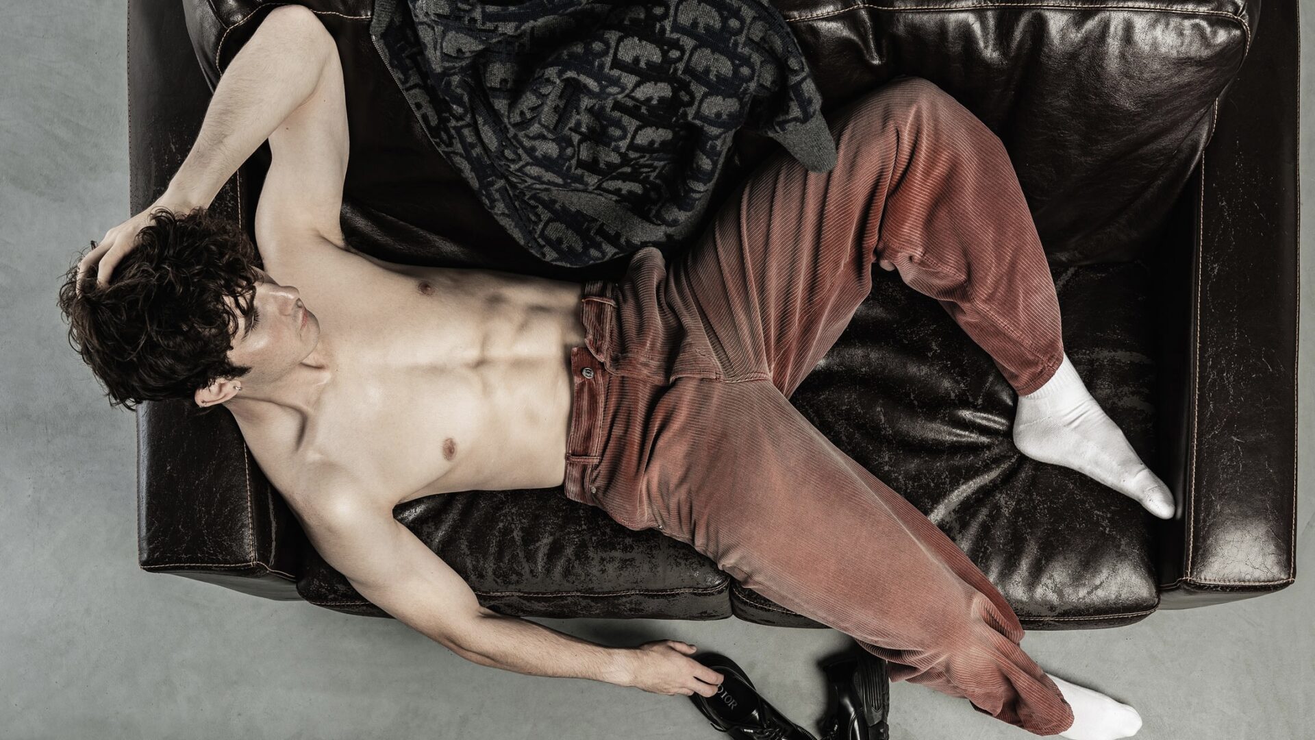 A model lays on a leather sofa wearing Dior Men's Burgundy trousers.