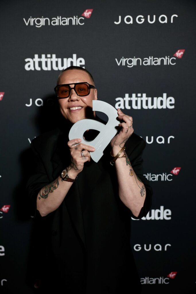 Gok Wan in black holding award, branding board behind him