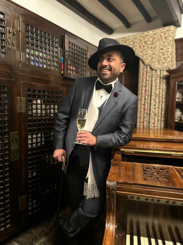 A man in a black suit and bow tow and hat holding a glass of champagne while leaning on a piano