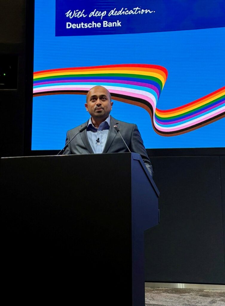 A man standing on stage at a lecturn with a rainbow flag on screen behind him
