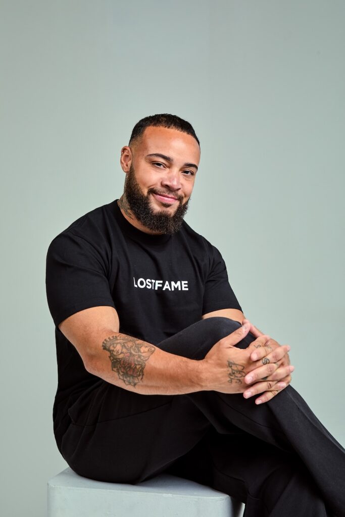 Kenny Ethan Jones in black sitting on a white stool, smiling, holding knee