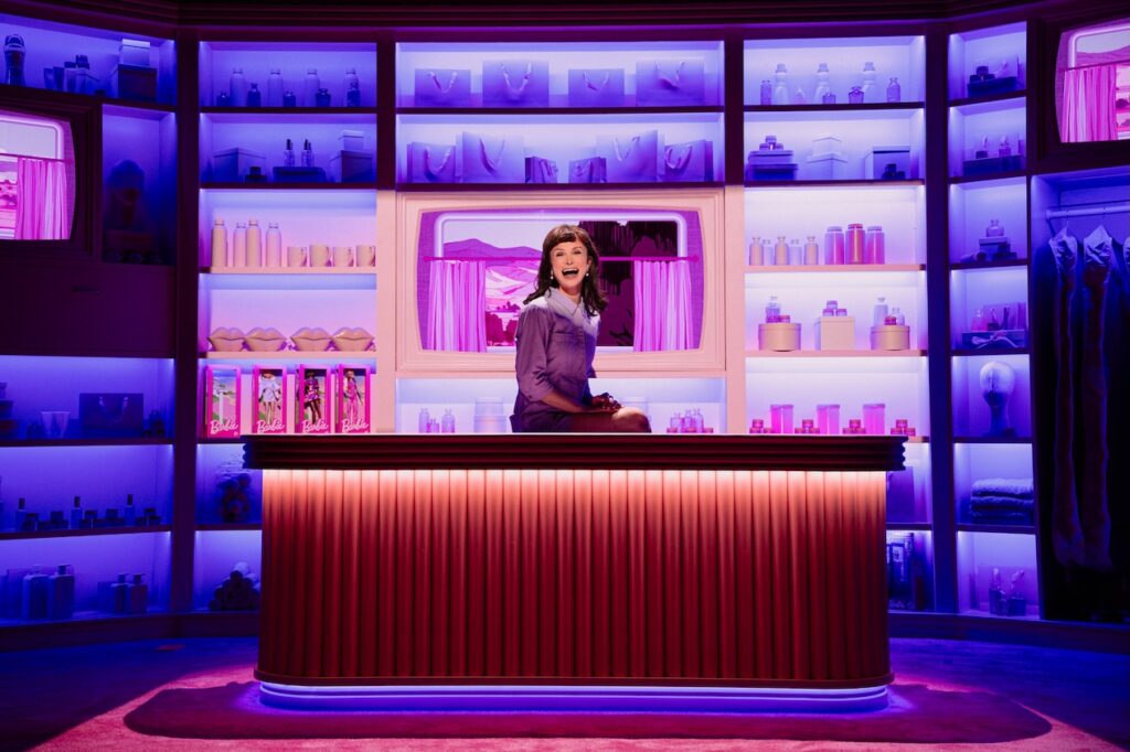 Dylan Mulvaney behind a counter on stage wearing a purple jacket
