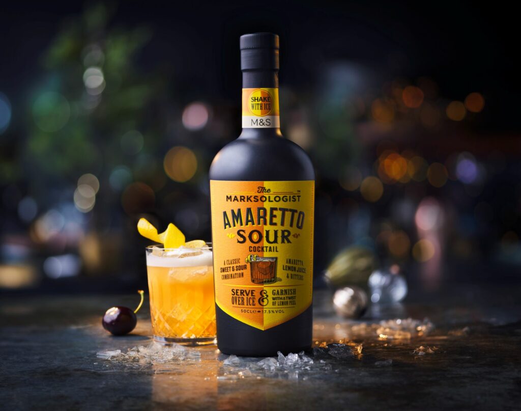 A bottle of M&S Marksologist Amaretto Sour next to a glass filled with orange liquid and a lemon peel