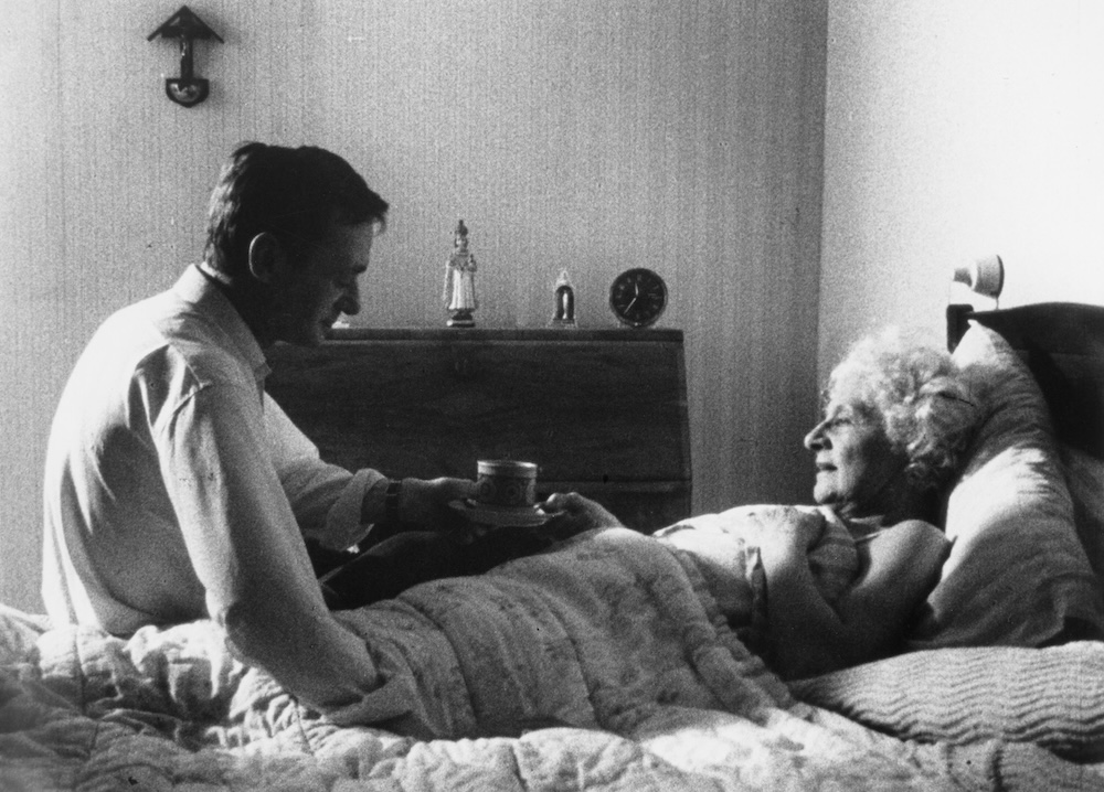 A still from Madonna and Child - a man handing an elderly lady a cup of tea as she lies in bed