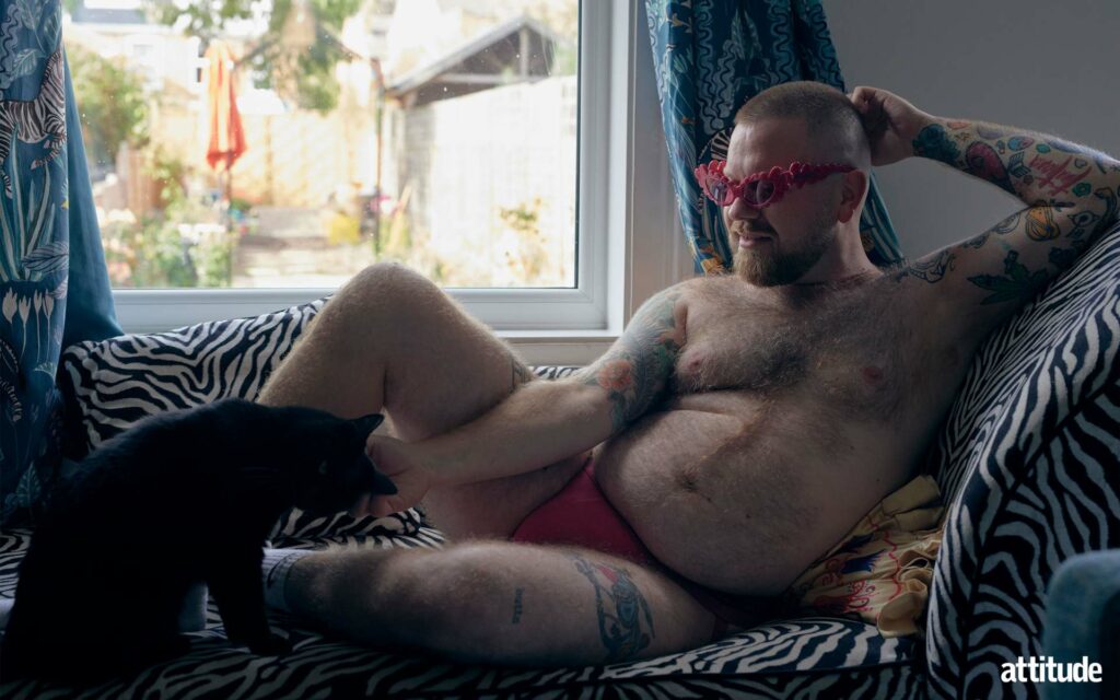 Ginny Lemon wearing pink underwear reclining on a sofa with a black cat