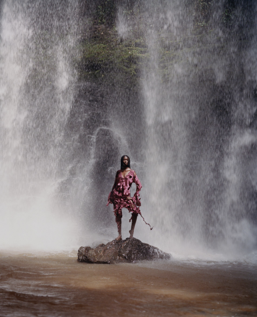 a model in a pink dress stands on a rock beneath a waterfall, photographed by Campbell Addy
