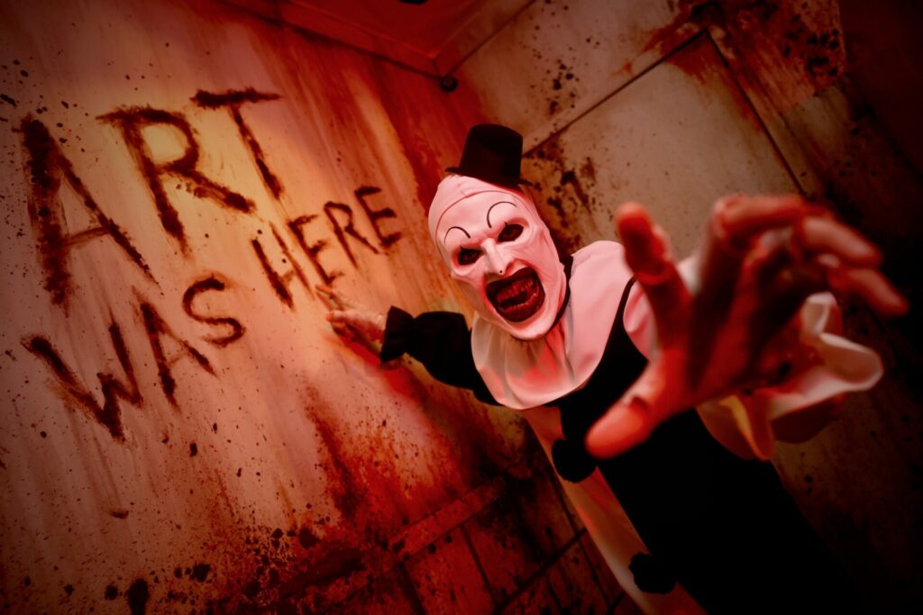 Art the Clown from Terrifier at Universal Horror Nights - black and white clown in blood-strewn room with "Art was here" written on wall in blood ((mage: Provided)