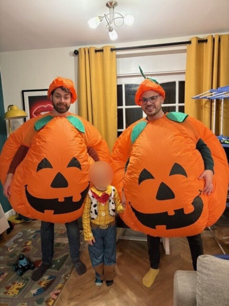 the dads in pumpkin outfits for halloween and their son, with face blurred, in a Woody from Toy Story costume