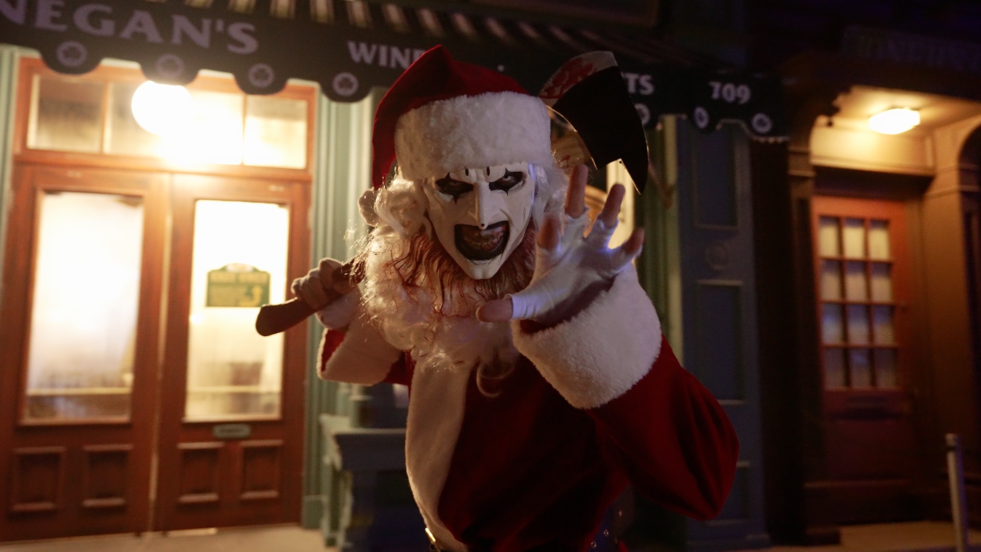 Art the Clown from the Terrifier films in a Santa outfit holding an axe outside a shop