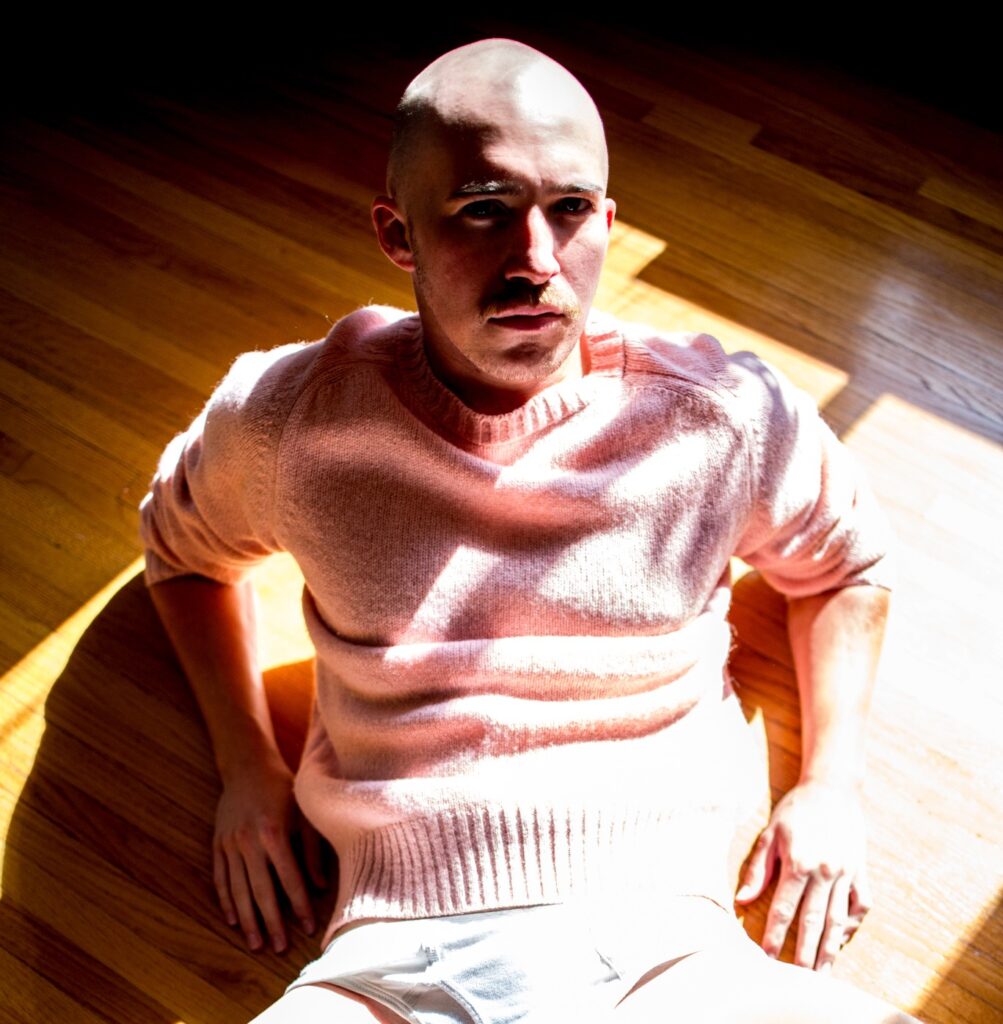 Model BB Gunn, in a pink jumper, lying on a wooden floor, shaved head and moustache