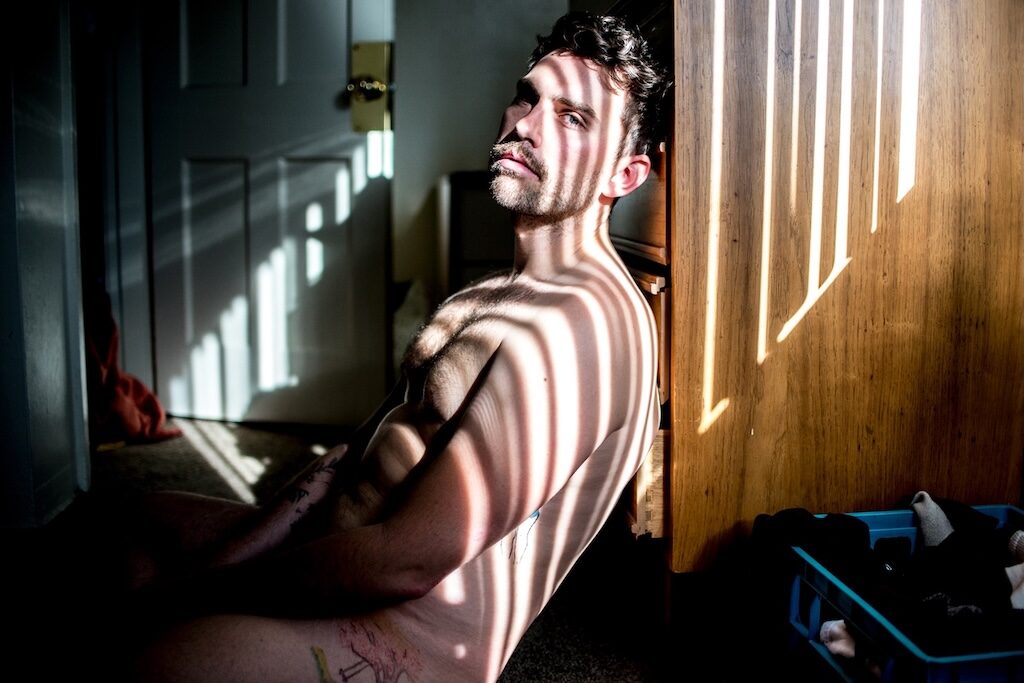 a model, Daniel Denhurt, with brown hair and moustache, sitting in a room with filtered sunlight over his face