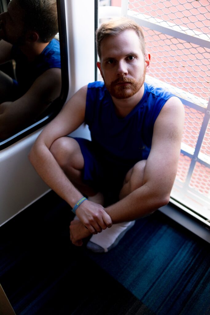 Matthew G, modelling for Elska, in a blue vest sitting on the floor in a train carriage