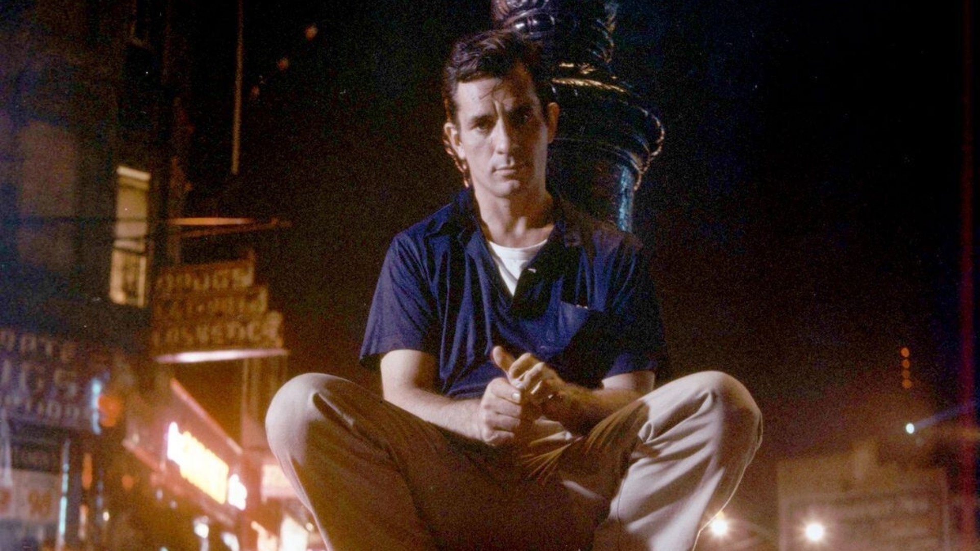 Jack Kerouac knelt down in blue shirt and white trousers, outdoors against a lamppost
