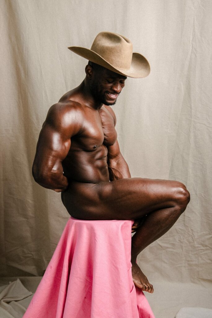 Ready to Ride’ portrait of Daniel Shoneye in a hat sat naked on a pink stool