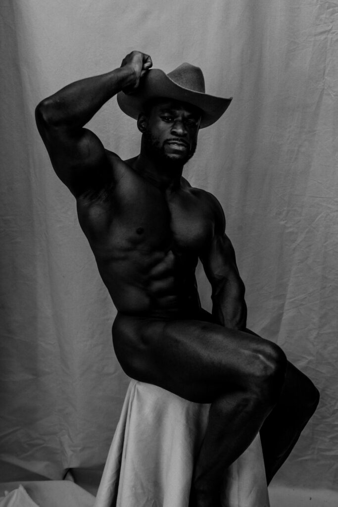 Ready to Ride’ portrait of Daniel Shoneye in a hat sat naked on a stool in black and white