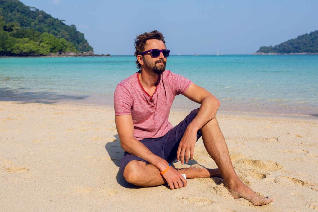 A smiling man in a pink T-shirt and blue shorts sitting on a beach with blue waters behind him