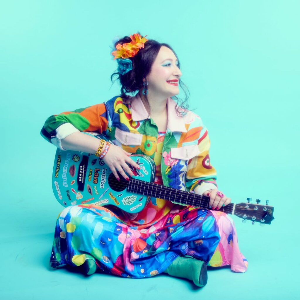 Rachael Sage dressed colourfully holding a blue guitar