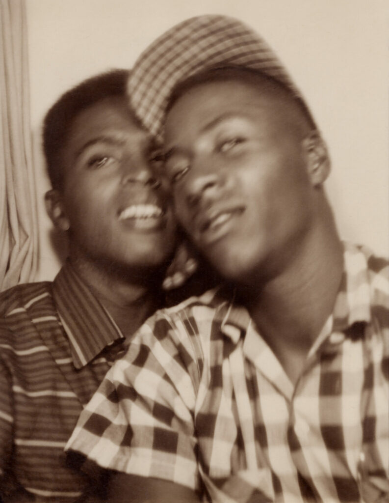a Black male couple in a Photo Booth smiling, one in a stripy t-shirt and one in a checked shirt