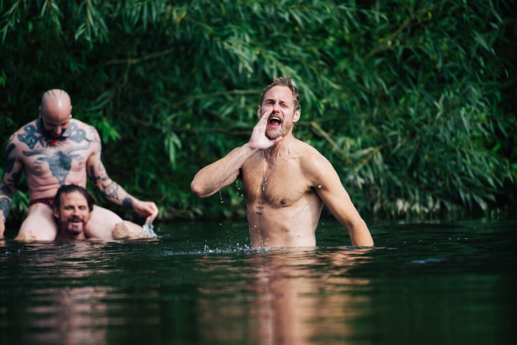 Alexander Skarsgård topless in a lake shouting towards the camera as two men look in; a still from the film Pillion
