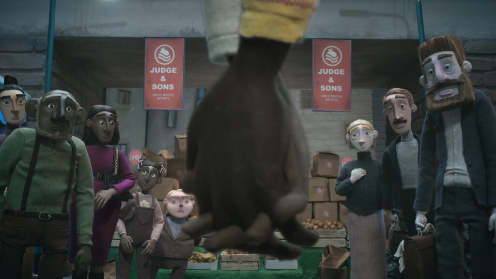 Dula and Edan’s hands are clasped in the foreground with a crowd watching in front of a fruit and
veg stall called ‘Judge & Sons.’