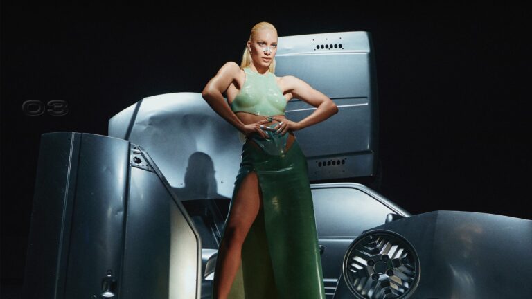 Agnes in a green dress with a car-like sculpture in the background, and a black wall