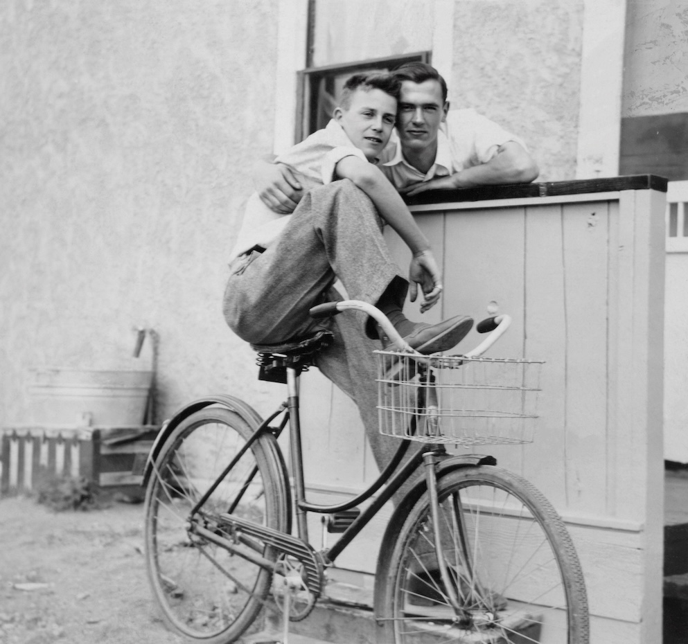 a male couple embracing in white shirts, one on a bicycle