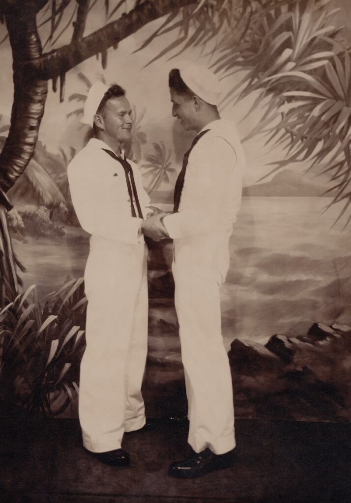 a male couple in sailors outfits looking at each other and holding hands, with a painted tree backdrop