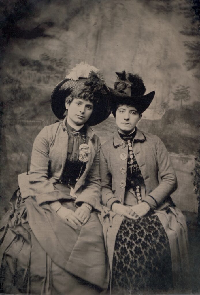 a female couple in hates, coats and dresses sitting side by side and looking at the camera