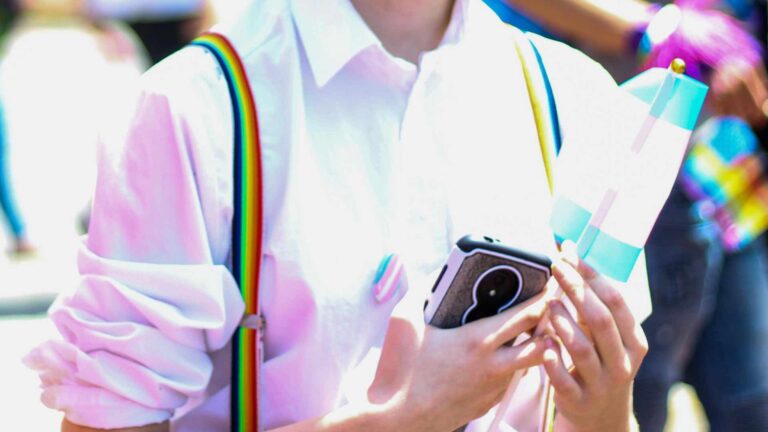 Someone holding a trans flag and wearing a trans pin badge holding their phone