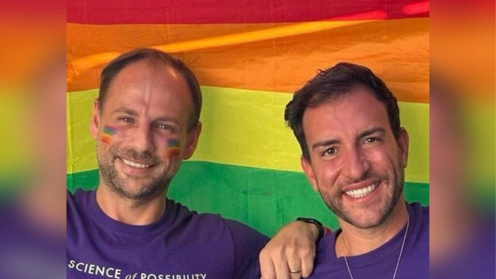 Two smiling men in purple T-shirts against a rainbow coloured backdrop