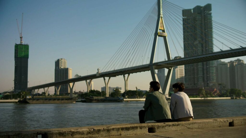 Two men sitting side by side in front of a bridge in Thai BL series Not Me