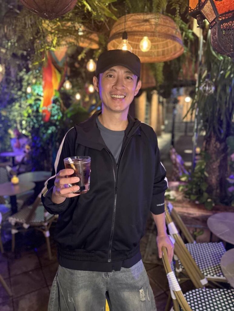 A smiling man in a black tracksuit jacket holding a glass of Coke
