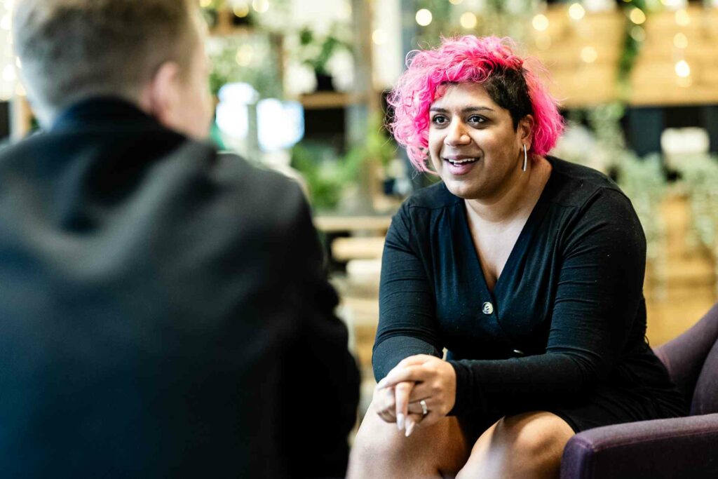 A person with pink hair sitting down chatting to another person
