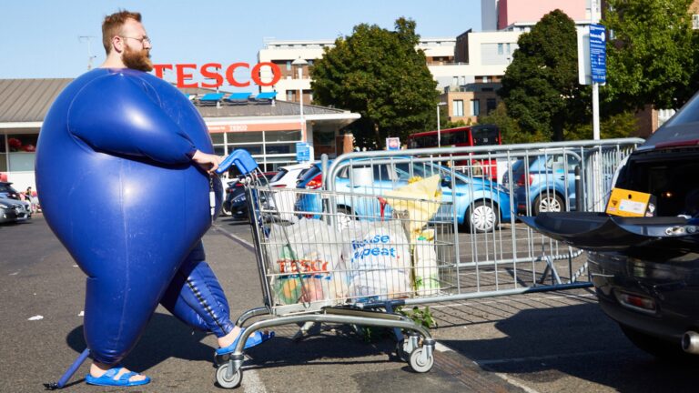 A model for Matt Ford pushes a shopping trolley in a Tesco's car p[ark while wearing an inflatable blue bodysuit