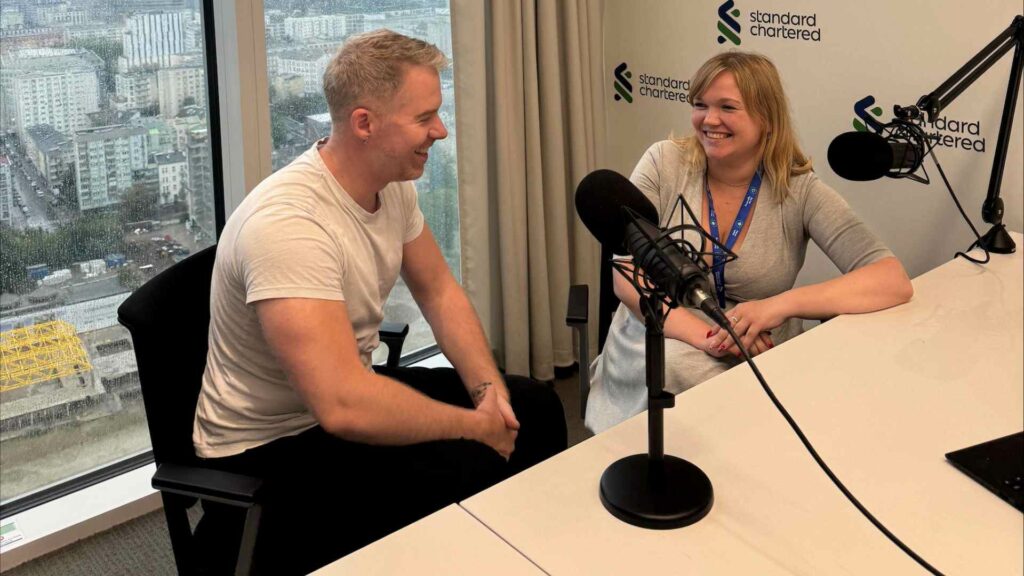 A man and woman talking in a podcast studio with a city backdrop behind them through a large window