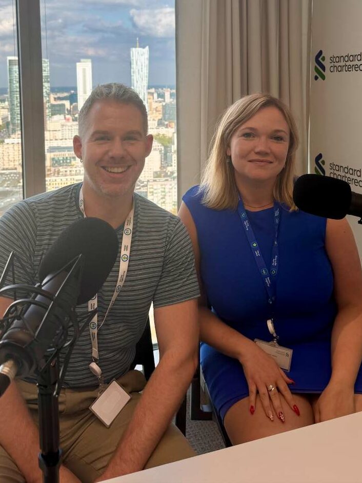 A man and woman smiling in a podcast studio with a city backdrop behind them through a large window