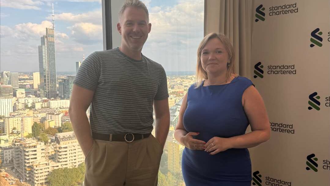 A man and woman standing side by side in front of a large window with a city view