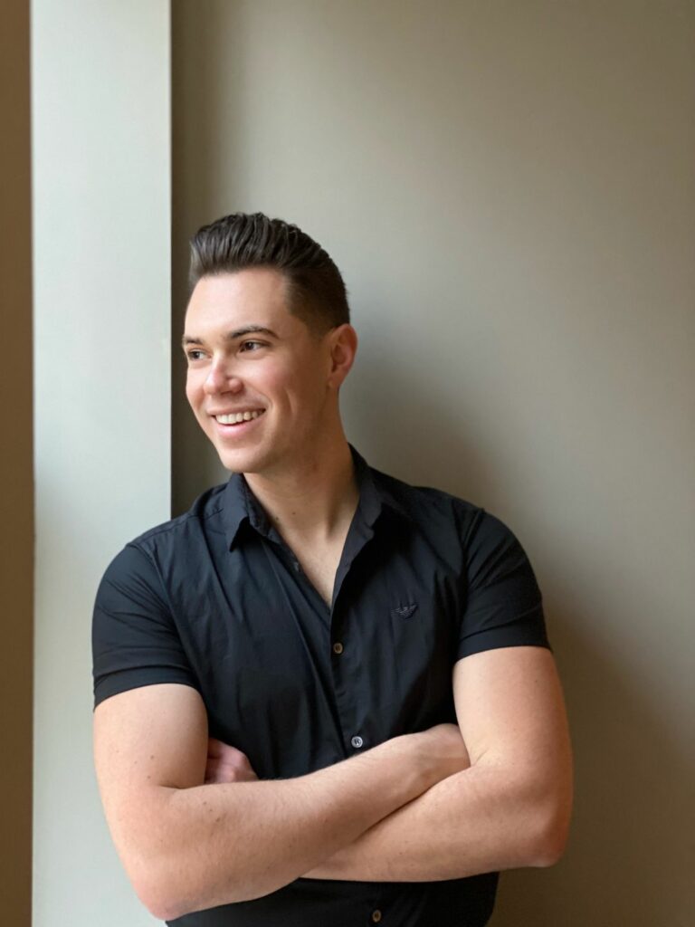 A smiling man with a short sleeved T-shirt folding his arms looking away from the camera