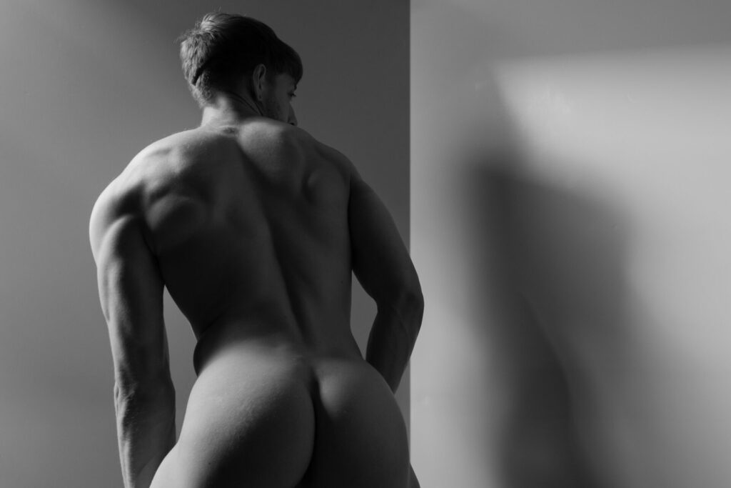 a photograph by Arturo Goni, showing a muscular nude man from behind in black and white