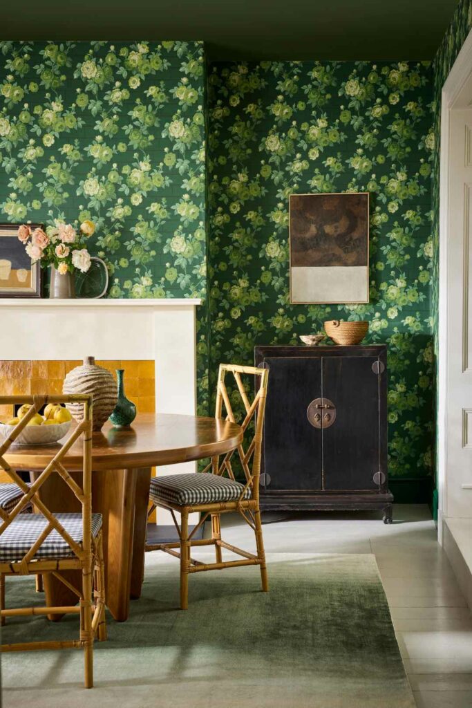Dining room with green floral wallpaper, wooden table and chairs, and a fireplace with decorative objects.