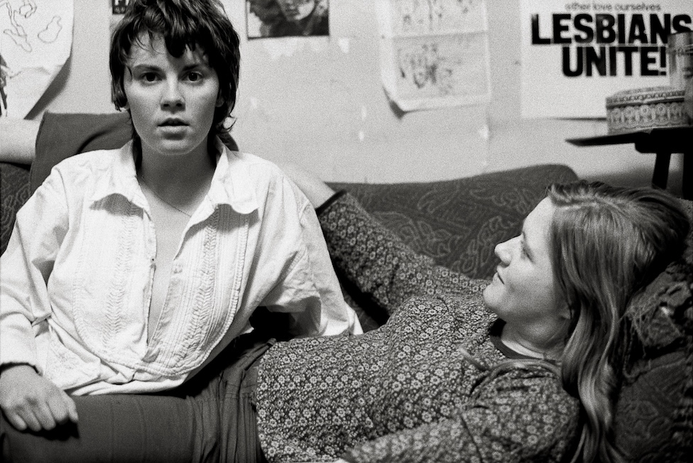 two people sitting/lying on a sofa, a poster says ';lesbians unite!' in the background