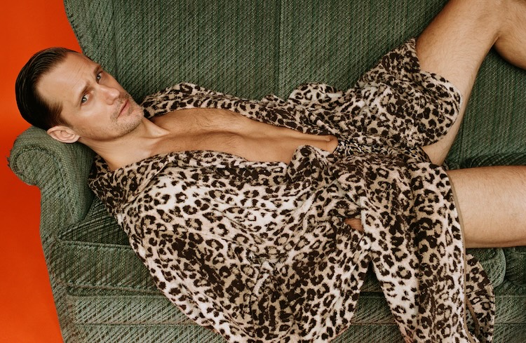 Alexander Skarsgård wearing a leopard print robe laying down on a green sofa