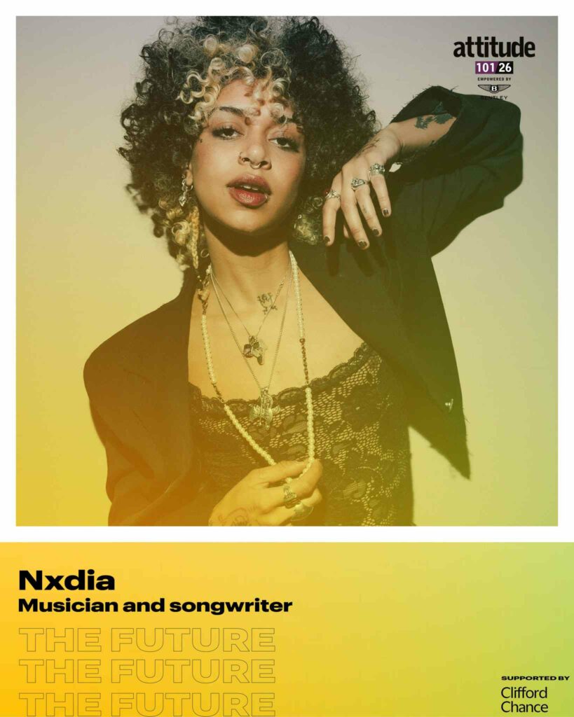 Nxdia posing in a black outfit with layered necklaces against a light background, with the Attitude 101 logo visible in the top corner.