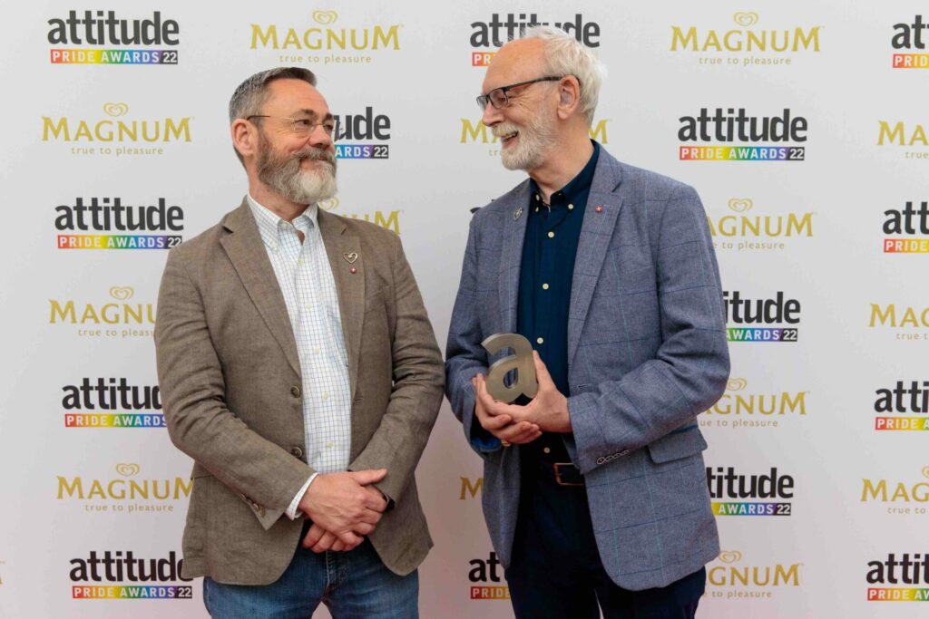 Rupert Whitaker and Martyn Butler OBE at the Attitude Pride Awards 2022 holding their award on the red carpet whilst looking at one another